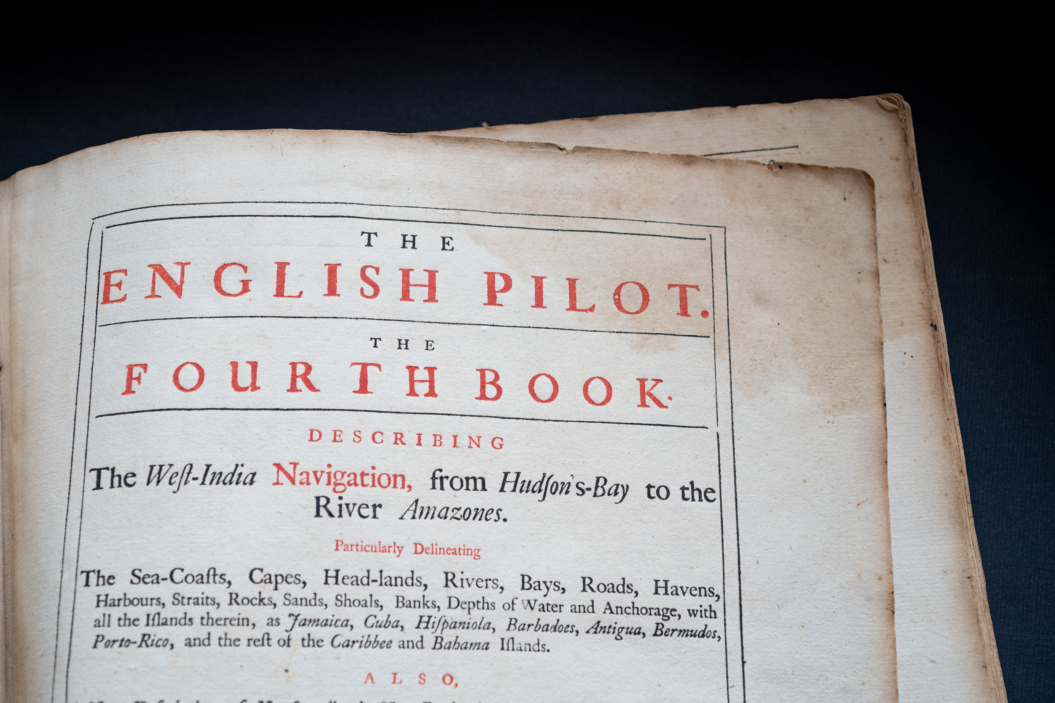 Detail of a printed book shows title page with English text "The English Pilot. The Fourth Book describing the West India Navigation, from Hudson's Bay to the River Amazones" in red ink.