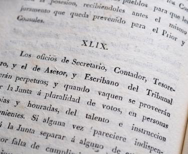 Detail of a printed book shows text in Spanish reading "Los oficios de secretario, Contador, tesorero" and so on.
