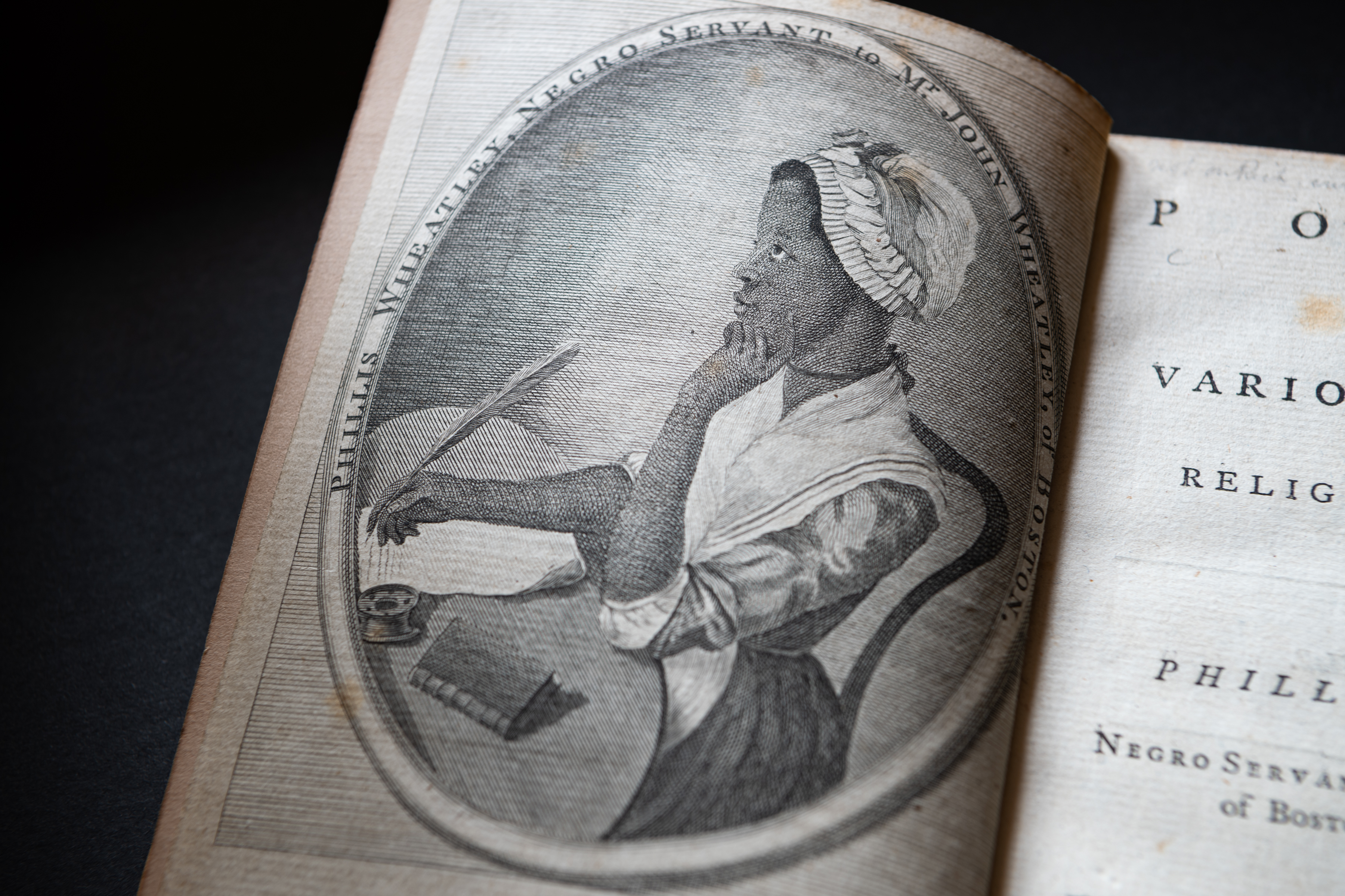 Detail of a printed book shows a portrait frontispiece of Phillis Wheatley sitting at a table with a quill in her hand in the process of writing.