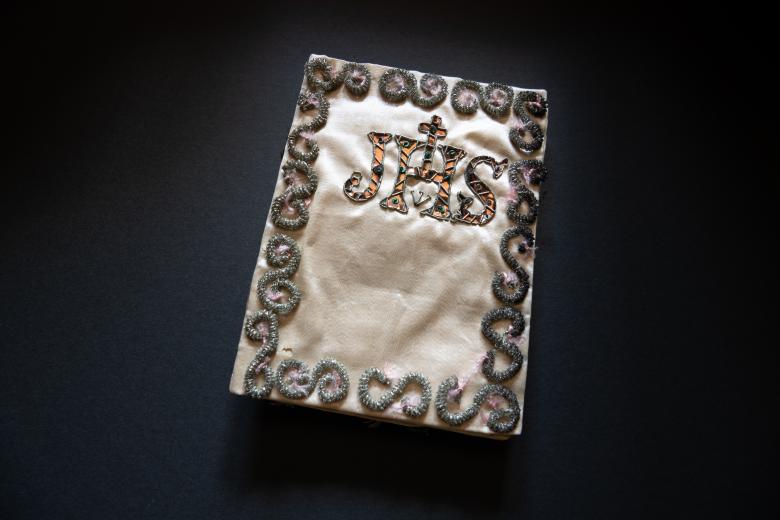 Book bound in tan silk bordered with beading and embroidered with letters "JHS" in the center.