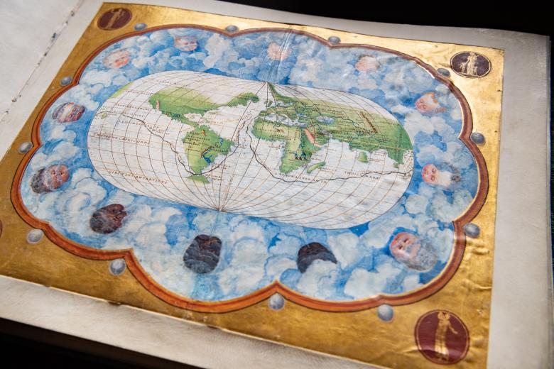 Detail of a colored manuscript map shows elliptical layout of the world. Wind heads, seen within clouds, surround the circumference of the map. Gilded border decorates the item.