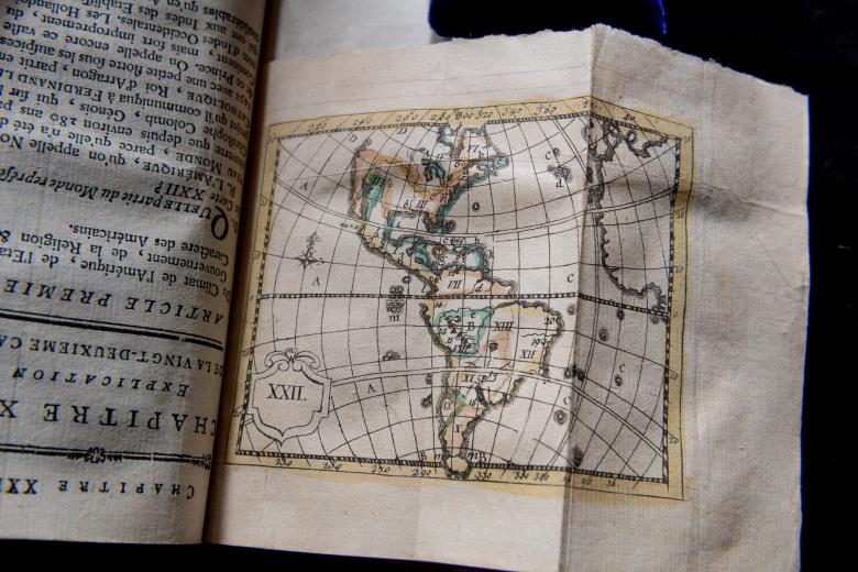 Detail of a printed book shows a fold-out map that includes latitude and longitude lines, orange and green colors, and roman numerals.