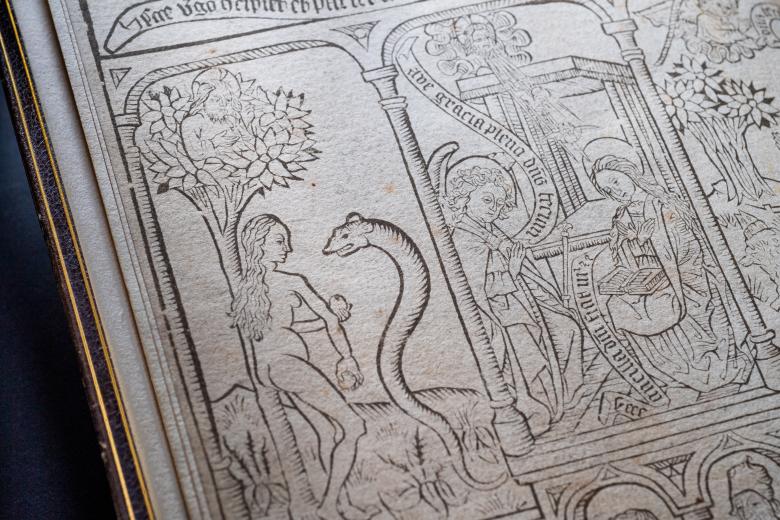 Detail of a blockbook shows woodcut illustration of biblical scene in the garden of eden. A woman holds an apple and is talking to a large snake. Text in Latin also visible.