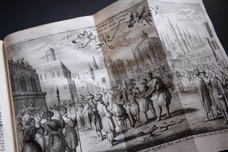 Detail of a printed book shows a fold-out illustration of people standing in a town square with buildings surrounding them and in the background. Text in Dutch.