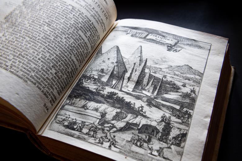 Detail of a printed book shows a full-page illustration of a mountainous landscape, including pyramid-like structures. Text in Dutch on the opposite page.