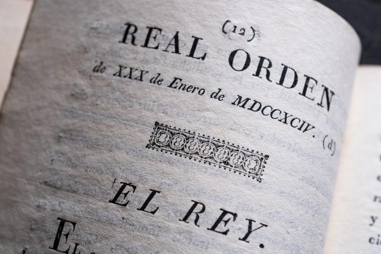 Detail of a printed book shows text in Spanish reading "Real Orden de XXX de Enero" and includes a decorative headpiece.