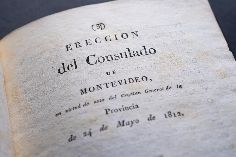 Detail of a printed book shows text in Spanish reading "Ereccion del consulado de Montevideo en virtud de auto del Capitan general de la provincia."
