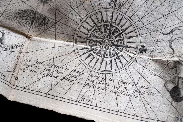 Detail of a printed fold-out map shows latitude and longitude lines, a large compass rose, decorative illustrations, and text in English.
