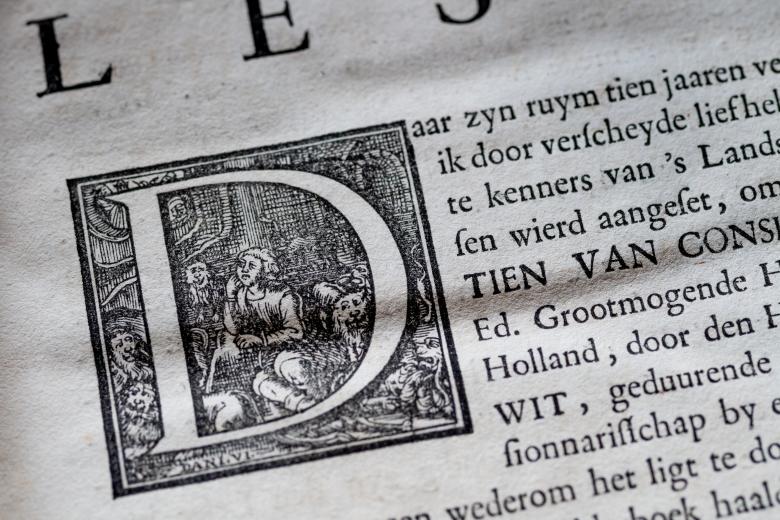Detail of a printed page shows the detailed typography as a "D" letter is seen as a block with people sitting in a room. Text in Dutch.