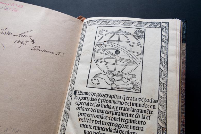 Detail of a printed book shows the title page with an illustration of a sphere and banners with title in Spanish "Suma de geographia..."