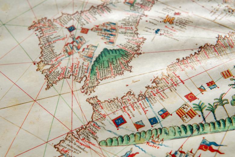 Detail of a colored manuscript atlas shows latitude and longitude lines, forests, and labels in Latin along a coastal region. Other details include castles and flags.