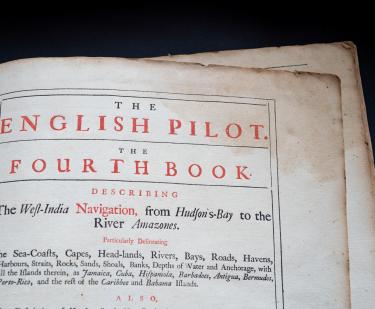 Detail of a printed book shows title page with English text "The English Pilot. The Fourth Book describing the West India Navigation, from Hudson's Bay to the River Amazones" in red ink.